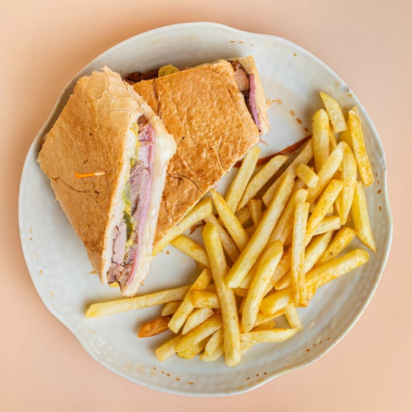 A pressed Cuban sandwich, cut in half, with a side of golden french fries on a white plate.